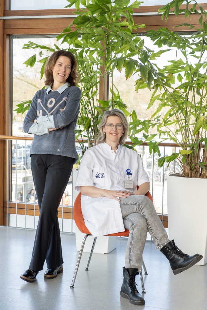 Twee dames poseren voor de camera, een met witte jas, kijken glimlachend in de camera